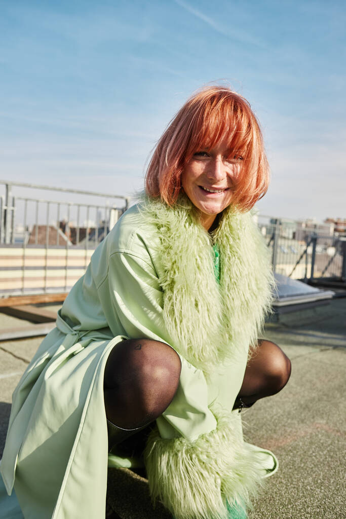 mujer alegre y de moda sentada en las horcas y mirando a la cámara en la terraza de la azotea en Viena - Foto, imagen