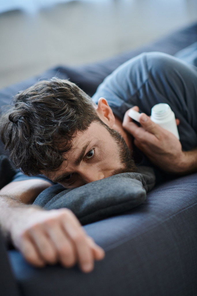 τραυματισμένος βασανισμένος άντρας με γενειάδα ξαπλωμένος σε καναπέ με χάπια στο χέρι, συνειδητοποίηση ψυχικής υγείας - Φωτογραφία, εικόνα