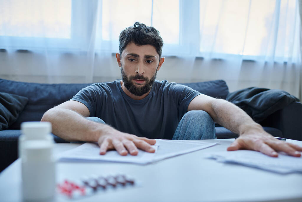 depressieve zieke man met baard zit aan tafel met papieren en pillen op het tijdens depressieve aflevering - Foto, afbeelding