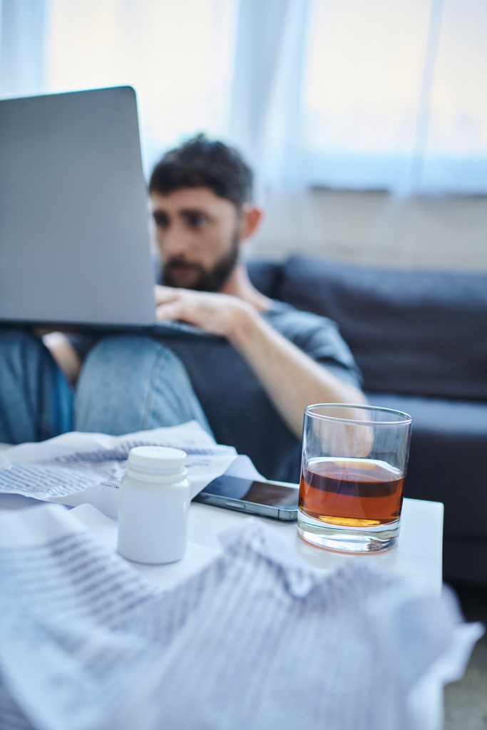 homme traumatisé déprimé avec barbe travaillant à l'ordinateur portable avec un verre de boisson alcoolisée sur la table - Photo, image