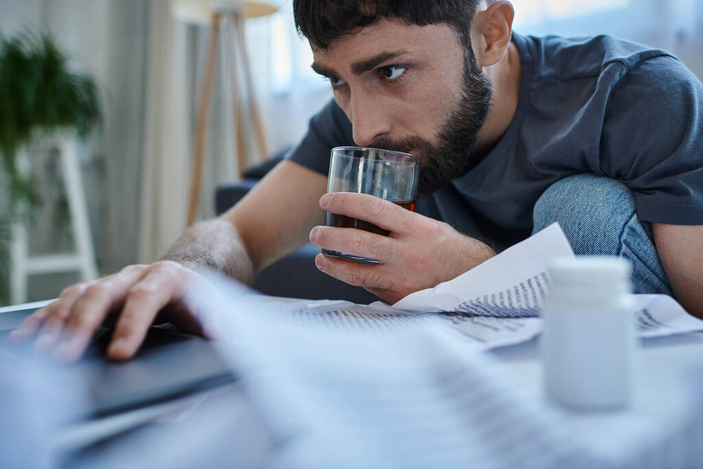 отчаянный травмированный человек с бородой работает за ноутбуком и пьет алкогольный напиток на столе - Фото, изображение