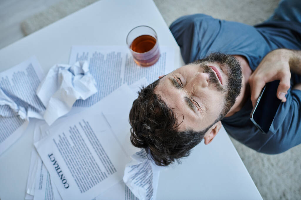 Zihinsel çöküntü sırasında yanında bir bardak alkol bulunan akıllı telefonlu depresif adamın üst görüntüsü. - Fotoğraf, Görsel