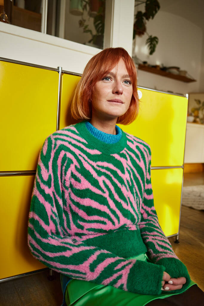 verträumte Frau im trendigen Animal-Print-Pullover sitzt in moderner Wohnung in Wien und schaut weg - Foto, Bild