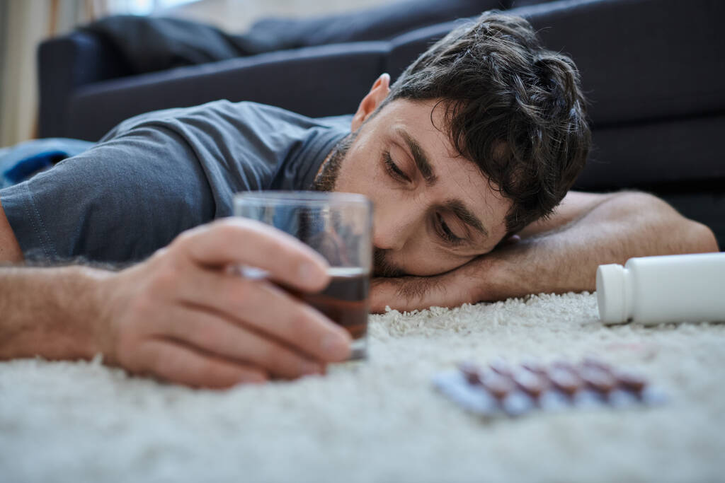 депрессивный человек в повседневной одежде пить алкоголь и смотреть на таблетки во время психического срыва - Фото, изображение