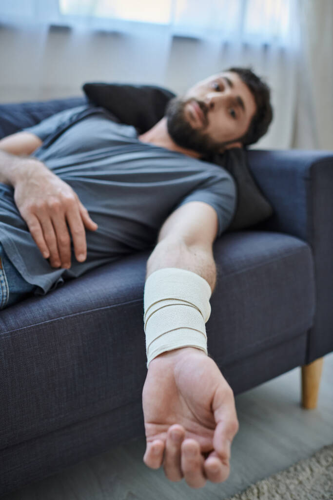 homme traumatisé avec bandage sur le bras après avoir tenté de se suicider couché sur le canapé, conscience de la santé mentale - Photo, image
