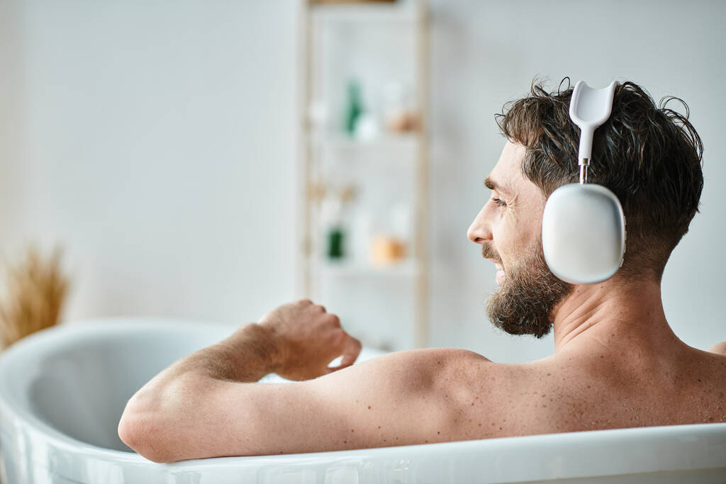 wesoły przystojny mężczyzna z brodą i słuchawkami siedzi i relaksuje się w wannie, zdrowie psychiczne - Zdjęcie, obraz