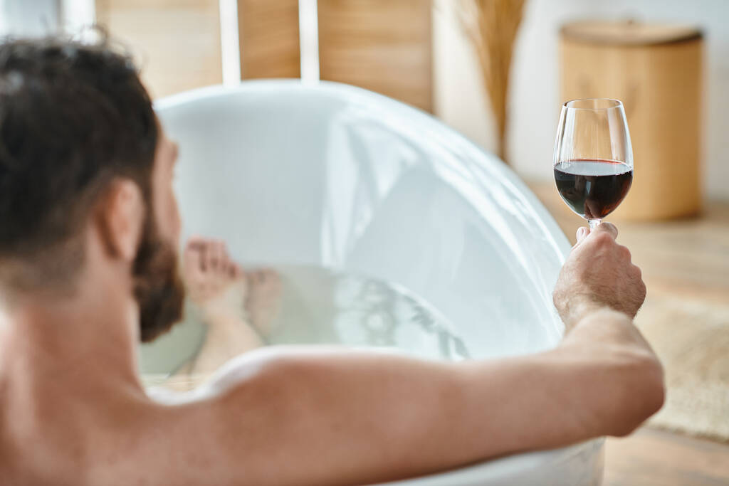 Сосредоточьтесь на бокале красного вина в руках размытого расслабляющего бородатого человека в ванной, психического здоровья - Фото, изображение