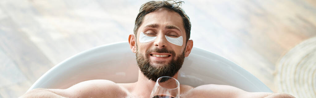 hombre atractivo alegre con barba y parches para los ojos relajante en la bañera con vaso de vino tinto, pancarta - Foto, Imagen