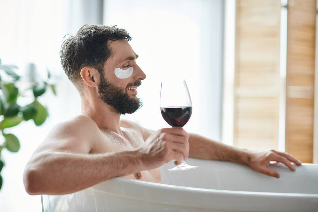 vidám jóképű férfi szakállal és szemtapasszal pihenés fürdőkádban egy pohár vörösborral - Fotó, kép