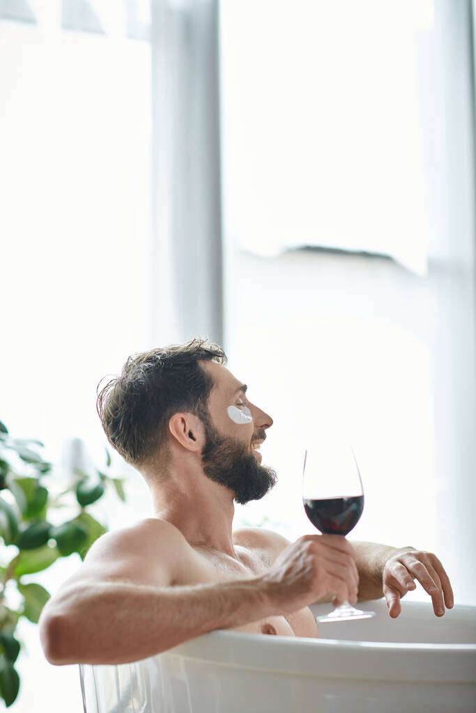 Sakallı ve göz bantlı neşeli yakışıklı adam elinde kırmızı şarapla küvette dinleniyor. - Fotoğraf, Görsel