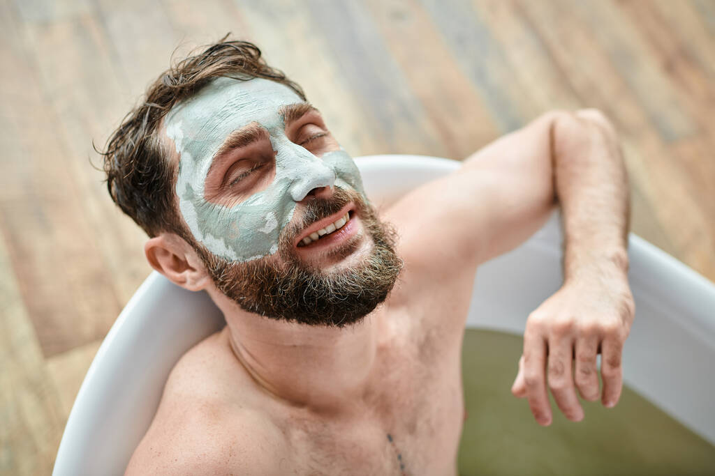 όμορφος χαρούμενος άντρας με γενειάδα και μάσκα προσώπου να παγώνει στην μπανιέρα του, συνειδητοποίηση ψυχικής υγείας - Φωτογραφία, εικόνα