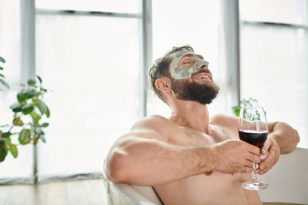 весёлый привлекательный мужчина с бородой и маской для лица расслабляется в ванной с бокалом красного вина - Фото, изображение