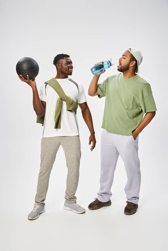 gelukkig afrikaanse amerikaanse man holding bal en kijken naar vriend drinken water op grijze achtergrond - Foto, afbeelding