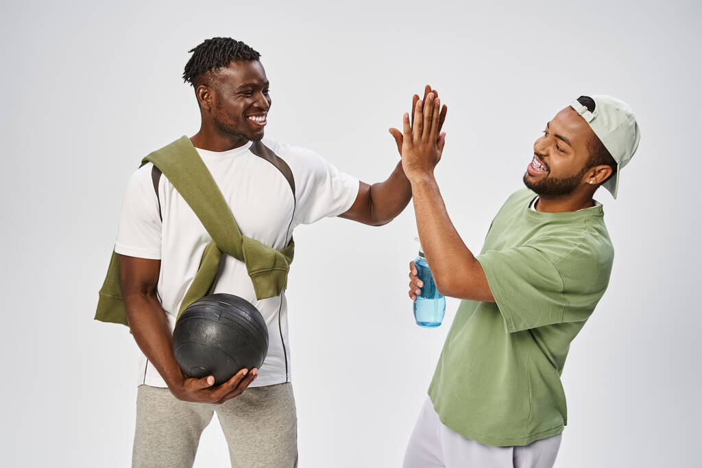 радісні афроамериканські друзі-чоловіки тримають м'яч і воду, даючи п'ять на сірому тлі - Фото, зображення