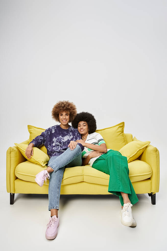 joyful and curly african american female friends sitting on yellow sofa on grey background - Photo, Image