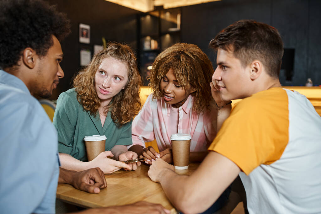 jonge en stijlvolle multi-etnische vrienden praten in de buurt van afhaaldrankjes in lobby cafe van studenten hostel - Foto, afbeelding
