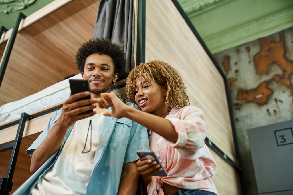 学生ホステルの部屋でスマートフォンを見ている若いアフリカ系アメリカ人のカップルの低い角度ビュー - 写真・画像