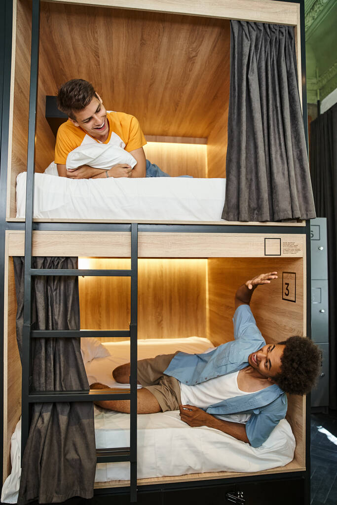 возбужденный африканский американский студент лежит на двухэтажной кровати и машет рукой другу в комнате общежития - Фото, изображение