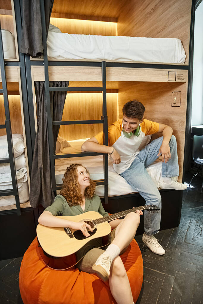 smiling woman playing acoustic guitar to boyfriend sitting on double-decker bed in hostel room - Photo, Image