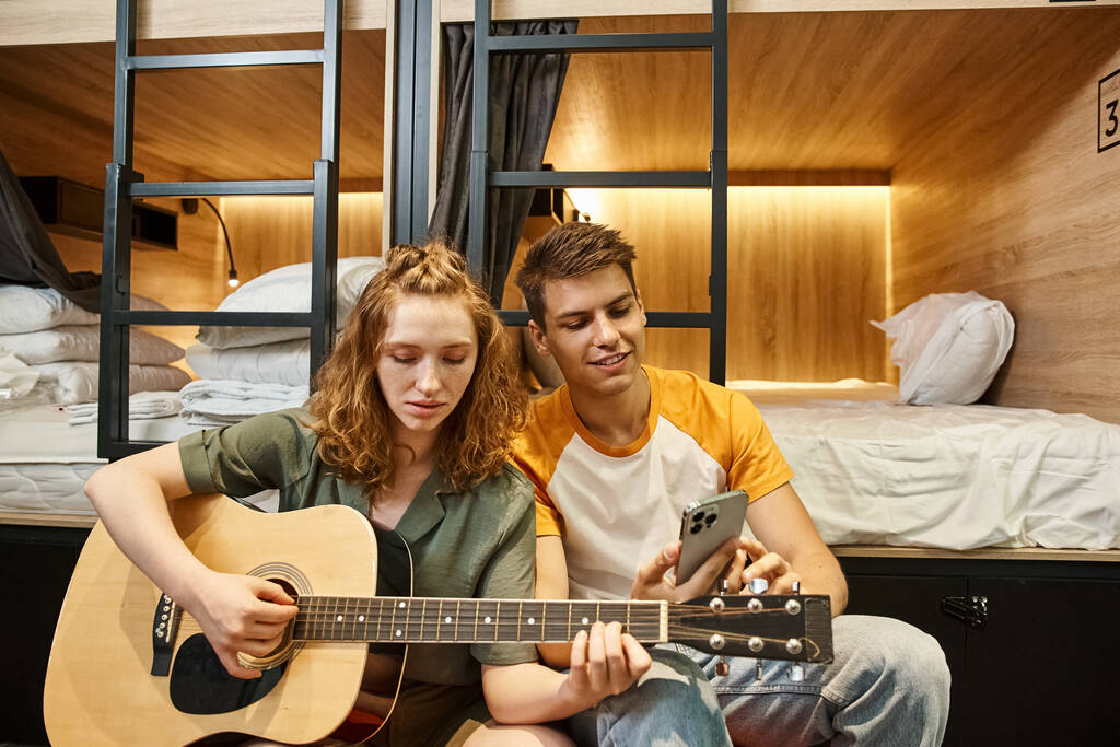 młoda kobieta gra na gitarze akustycznej do chłopaka ze smartfonem w przytulnym pokoju hostelu studentów - Zdjęcie, obraz