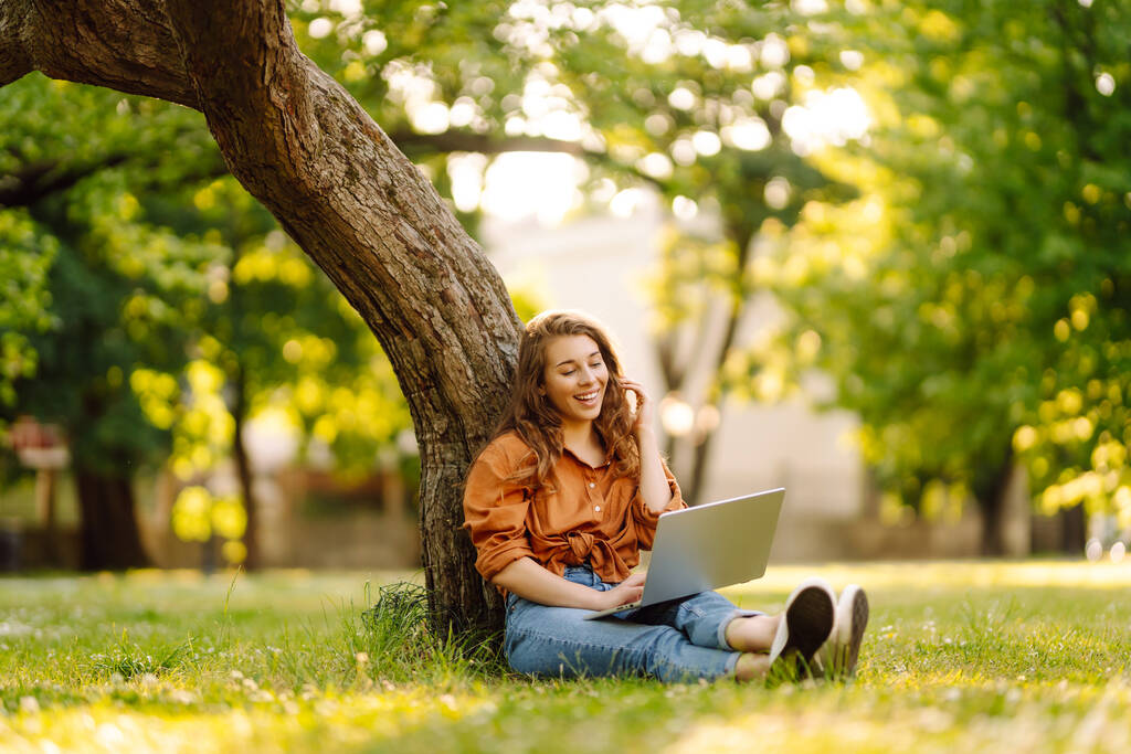 Mujer linda pasar tiempo al aire libre con el ordenador portátil. Mujer alegre trabaja en línea en el césped en el parque. Concepto de freelancing, caminar. - Foto, imagen
