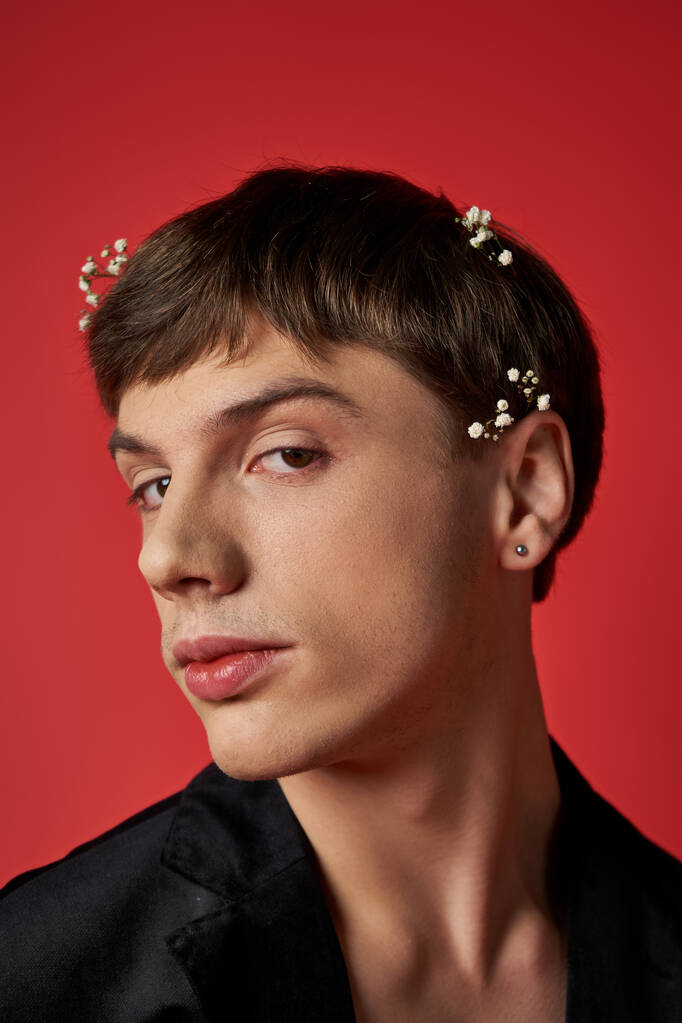 όμορφος νεαρός άνδρας με κομψό ντύσιμο με λουλούδια στα μαλλιά κοιτάζοντας κάμερα σε κόκκινο φόντο - Φωτογραφία, εικόνα