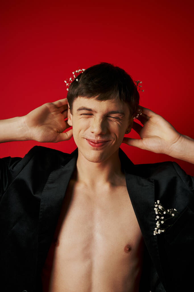 glücklicher Mann im eleganten Samt-Blazer mit Blumen im Haar lächelnd mit geschlossenen Augen auf rotem Hintergrund - Foto, Bild