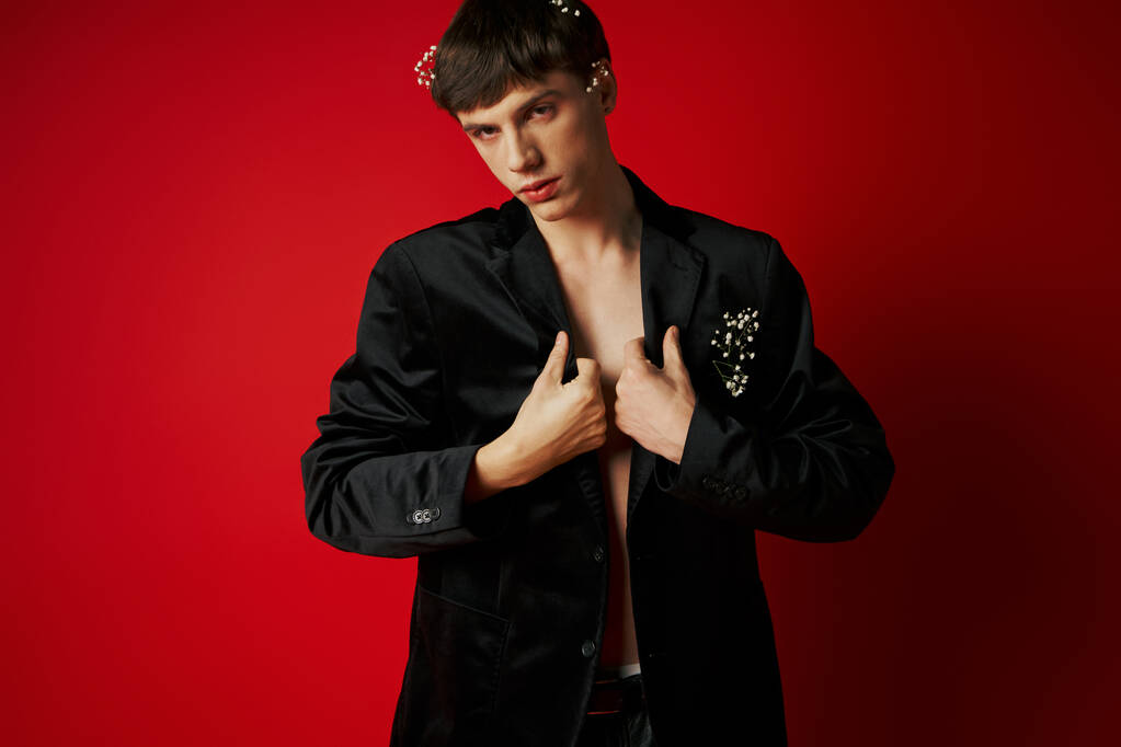 elegant young man in velvet blazer with flowers in hair looking at camera and posing on red backdrop - Photo, Image