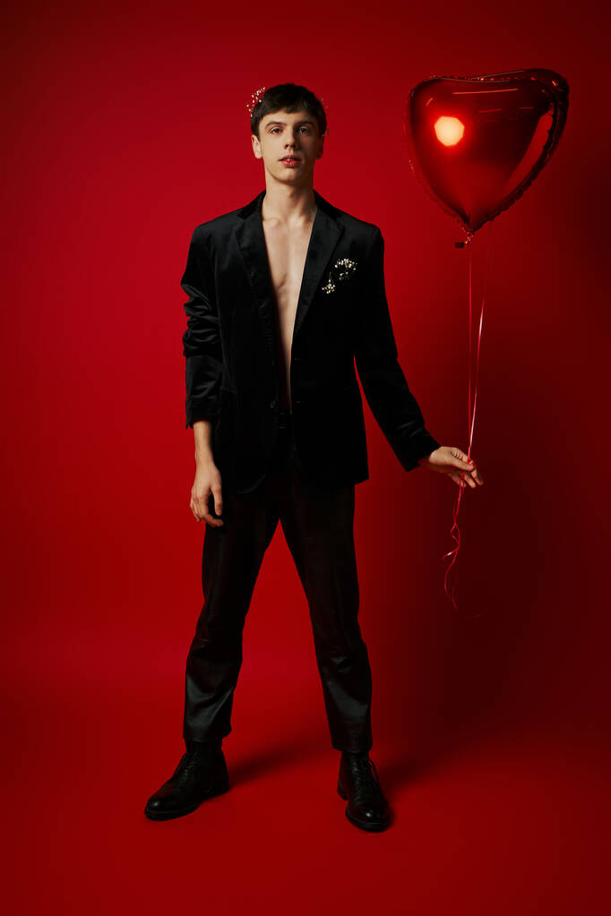 teljes hossza jóképű fiatal úriember fekete öltözetben tartja szív alakú léggömb piros háttér - Fotó, kép