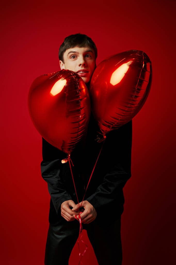 όμορφος νεαρός κύριος με μαύρη ενδυμασία κρατώντας μπαλόνια σε σχήμα καρδιάς σε κόκκινο φόντο - Φωτογραφία, εικόνα