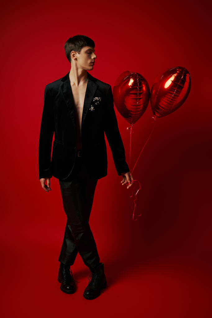 pleine longueur de beau monsieur en tenue noire tenant des ballons en forme de coeur sur fond rouge - Photo, image