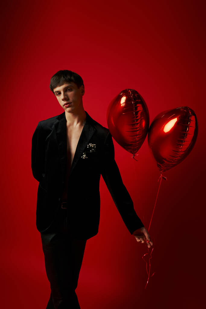 jong mannelijk model in fluwelen jasje en leren broek met hartvormige ballonnen op rode achtergrond - Foto, afbeelding