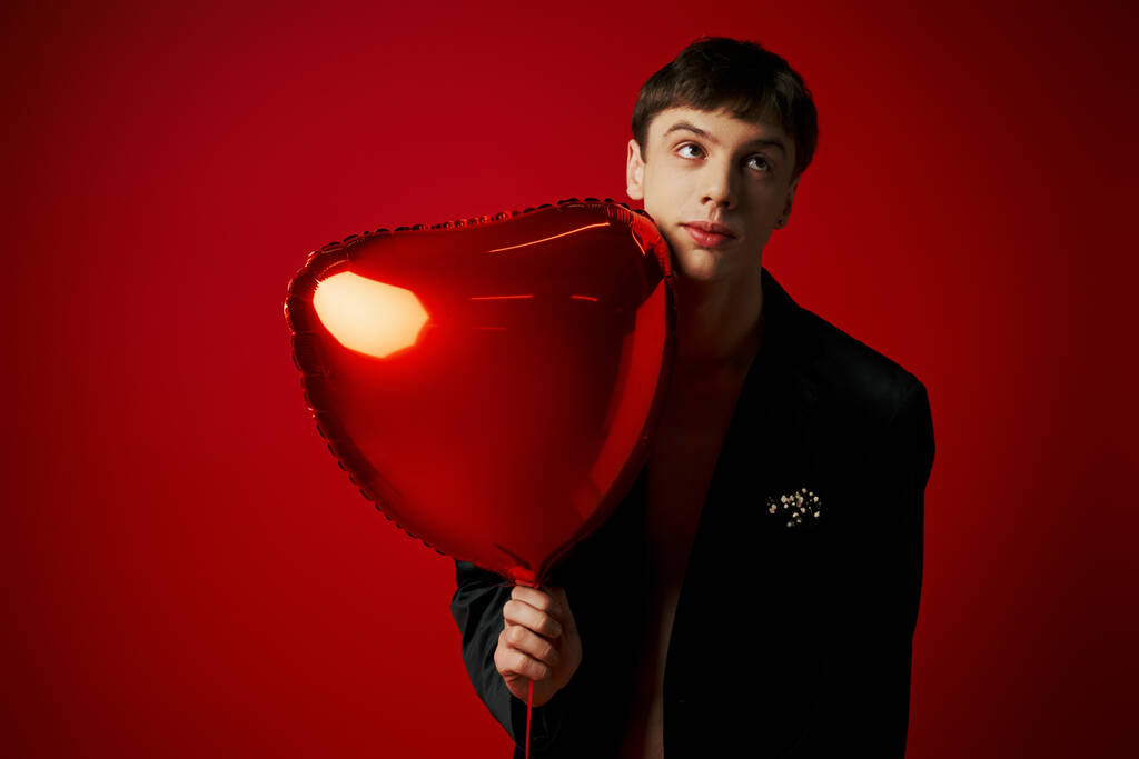 zdezorientowany młody człowiek w aksamitnej kurtce trzymając balon w kształcie serca i odwracając wzrok na czerwonym tle - Zdjęcie, obraz