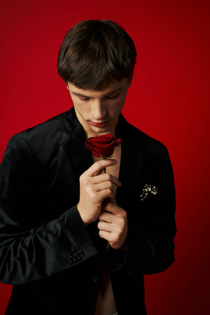 романтичный и элегантный молодой человек в бархатной куртке держа розу в руках на красном фоне, флирт - Фото, изображение