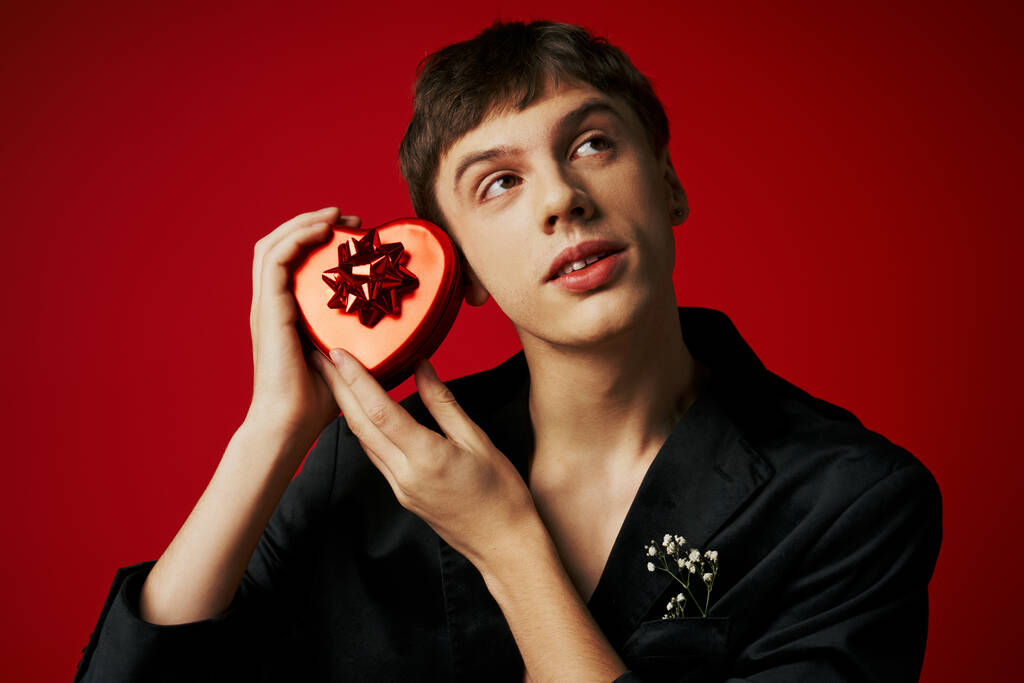 dreamy young man in velvet blazer holding heart-shaped present on red background, Valentines day - Photo, Image