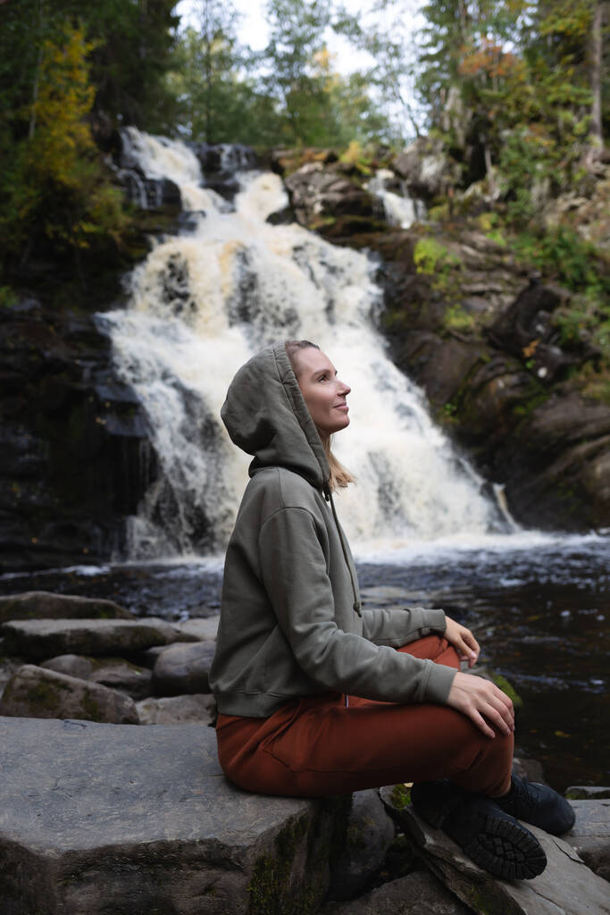 молода красива жінка-туристка, що сидить на тлі водоспаду, оточеного лісом. - Фото, зображення