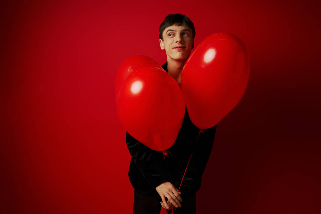 happy young man in black attire holding heart-shaped balloons on red background, Valentines day - Photo, Image