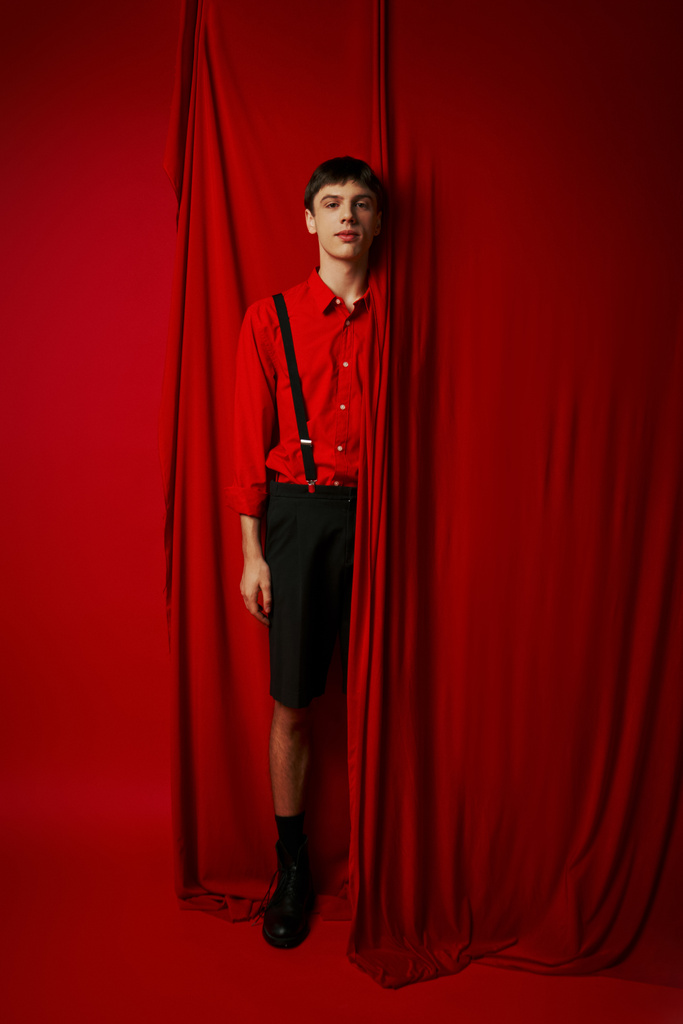 full length of young man in vibrant shirt with suspenders hiding behind red curtain, trendy look - Photo, Image
