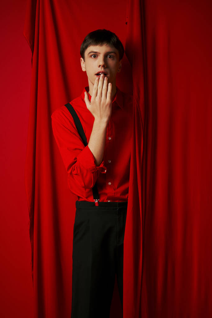 surprised young man in suspenders smiling and covering mouth near red vibrant curtain, merriment - Photo, Image