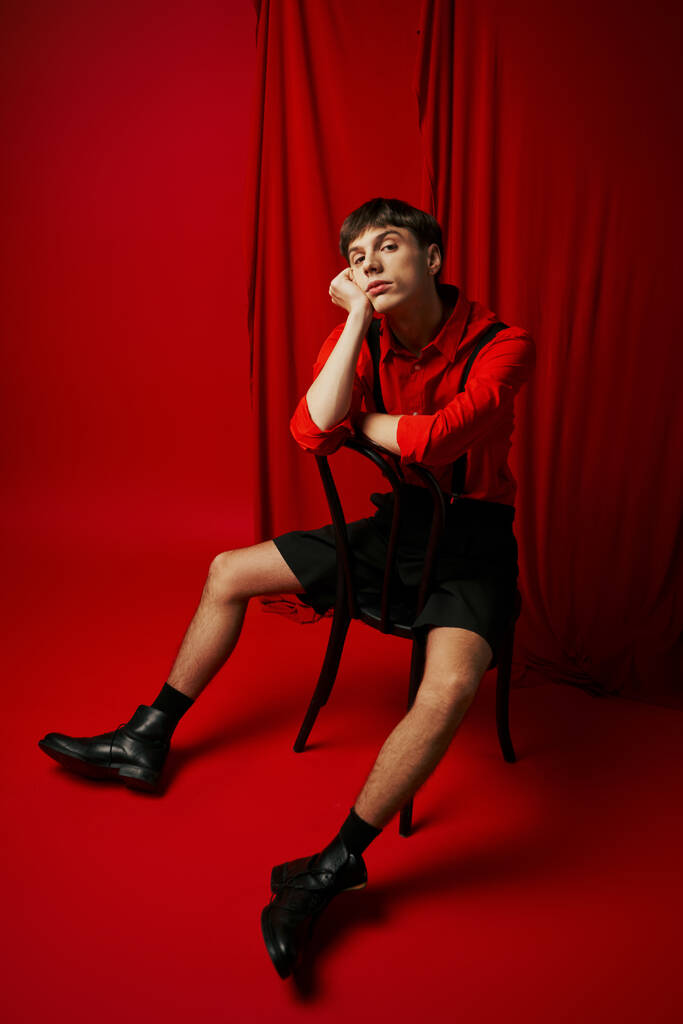 verveelde jongeman in hemd en zwarte short zittend op stoel met relaxte pose naast rood gordijn - Foto, afbeelding
