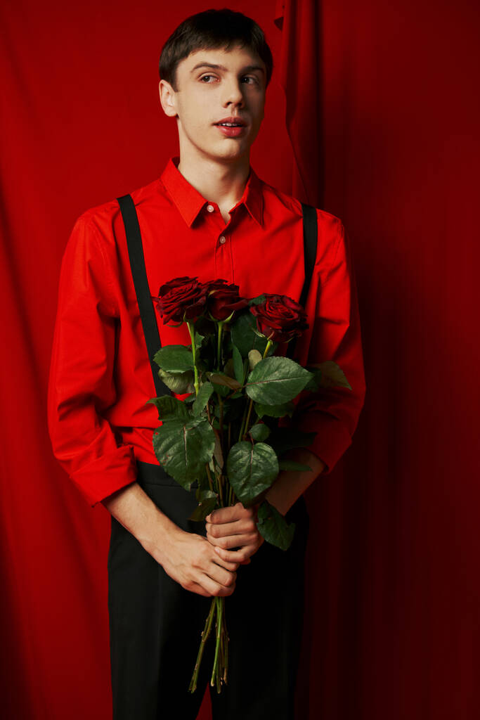 νεαρός άνδρας σε ζωντανή πουκάμισο και σορτς με τιράντες κρατώντας μπουκέτο τριαντάφυλλα κοντά στην κόκκινη κουρτίνα - Φωτογραφία, εικόνα