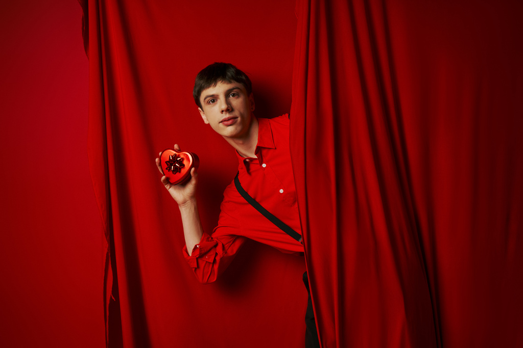 joven de camisa roja con tirantes sosteniendo caja de regalo en forma de corazón cerca de la cortina, 14 de febrero - Foto, Imagen
