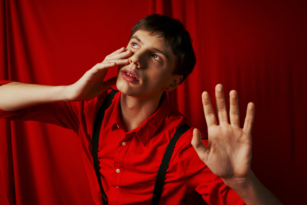 napínavý muž v košili s kšandami pózující se zdviženýma rukama na červeném pozadí, polekaný - Fotografie, Obrázek