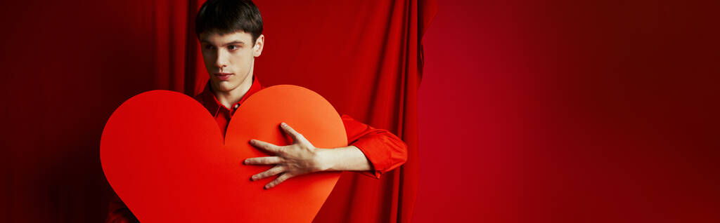 jeune homme en chemise embrassant grand carton en forme de coeur sur fond rouge, bannière Saint-Valentin - Photo, image