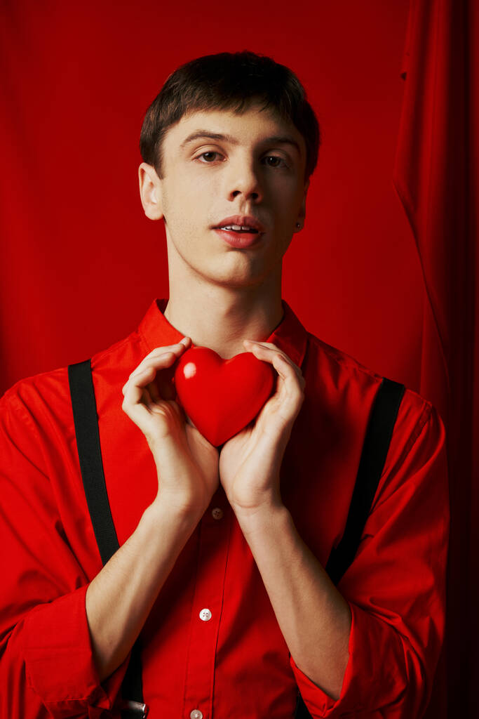νεαρός άνδρας κρατώντας μικρή καρδιά τρυφερά στα χέρια του και κοιτάζοντας κάμερα στο κόκκινο φόντο - Φωτογραφία, εικόνα