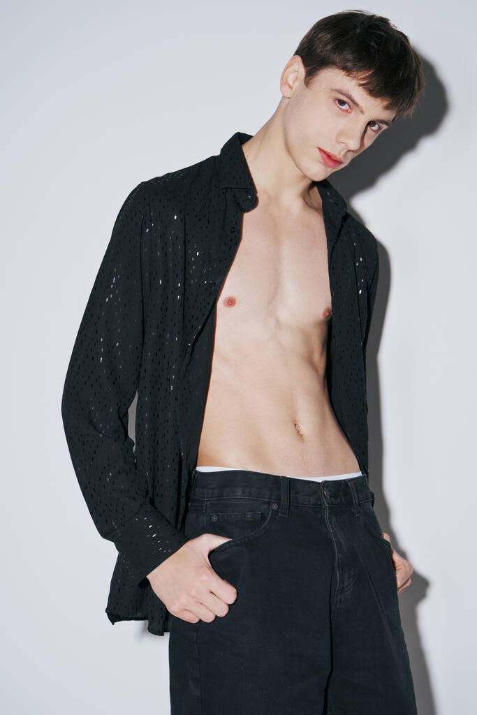 confiant jeune homme en chemise ouverte noire brillante avec doigt dans la poche de pantalon sur fond gris - Photo, image