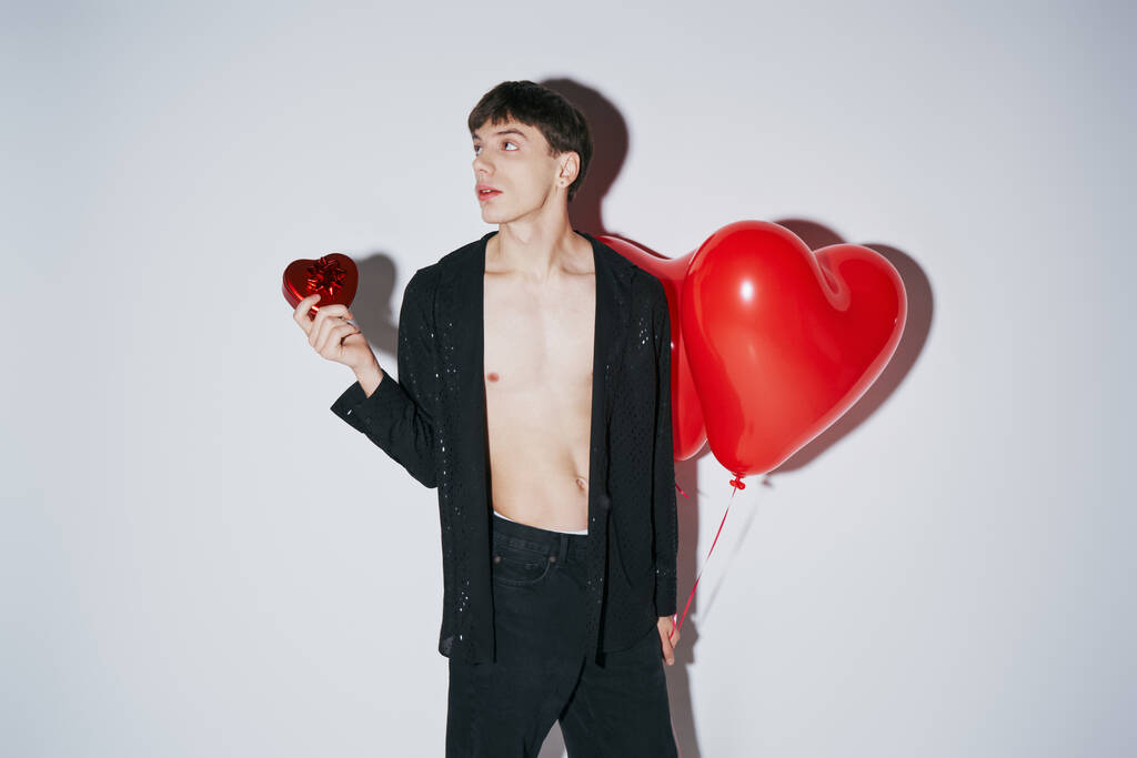Siyah gömlekli adam kırmızı kalp şeklinde balon tutuyor ve gri arka planda sevgililer gününde bulunuyor. - Fotoğraf, Görsel