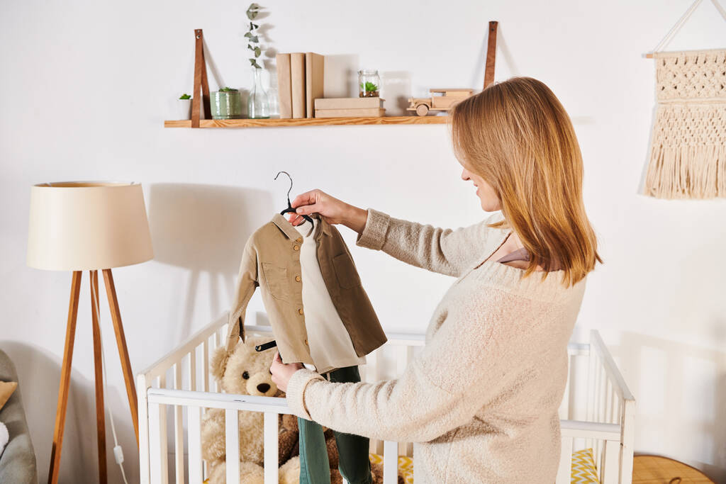 jonge toekomstige moeder houden babykleertjes in de buurt van wieg met zacht speelgoed in de kinderkamer, geboorteverwachting - Foto, afbeelding