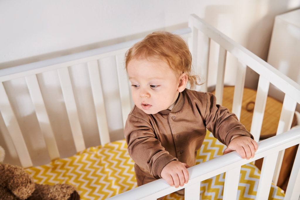 vysoký úhel pohledu na rozkošné dítě v rampě stojící v postýlce v dětském pokoji doma, batole - Fotografie, Obrázek