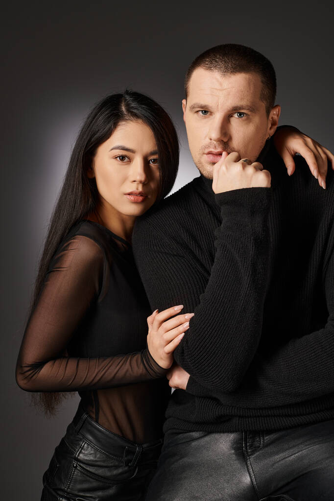 fashionable expressive interracial couple in black clothes looking at camera on grey backdrop - Photo, Image
